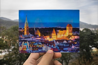 A postcard being held in front of a blurred outdoor background. The postcard features a vibrant scene of a cityscape at dusk, highlighting illuminated buildings, including a prominent church with a tall spire and other historical architecture. The text on the postcard reads 'Toruń, Poland.'