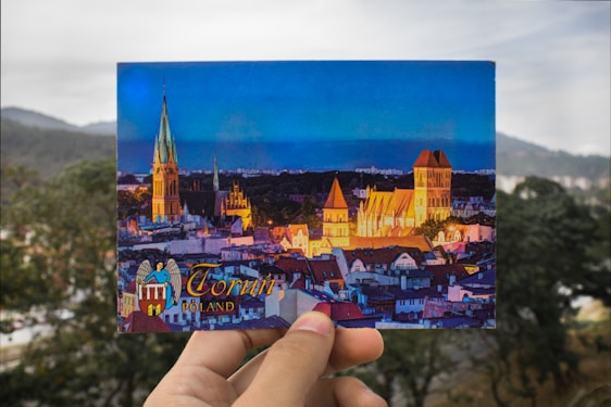 A postcard being held in front of a blurred outdoor background. The postcard features a vibrant scene of a cityscape at dusk, highlighting illuminated buildings, including a prominent church with a tall spire and other historical architecture. The text on the postcard reads 'Toruń, Poland.'