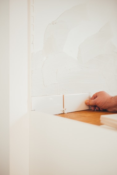 A person is installing white ceramic tiles on a wall, using a spacer to ensure even gaps. The wall has adhesive spread over it in preparation for tiling, with a wooden surface at the bottom edge.