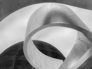 A large metallic sculpture with scientific formulas and text is prominently displayed against a cloudy sky. The structure appears to be twisted and curved, giving it a dynamic shape, while a dome-shaped building is visible in the background. The texts and symbols indicate a focus on scientific and mathematical themes.