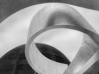 A large metallic sculpture with scientific formulas and text is prominently displayed against a cloudy sky. The structure appears to be twisted and curved, giving it a dynamic shape, while a dome-shaped building is visible in the background. The texts and symbols indicate a focus on scientific and mathematical themes.