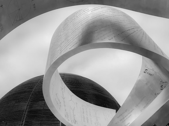A large metallic sculpture with scientific formulas and text is prominently displayed against a cloudy sky. The structure appears to be twisted and curved, giving it a dynamic shape, while a dome-shaped building is visible in the background. The texts and symbols indicate a focus on scientific and mathematical themes.