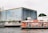 A modern building with a reflective glass facade features prominently, showing the words 'River City Bangkok' and promoting itself as an 'Anchor of Arts and Antiques.' In front of the building, a red and white ferry is docked, with passengers visible onboard. The setting is alongside a river and includes additional signage with local language script. Lush greenery decorates the area near the entrance.