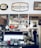 A cozy cafe interior featuring vintage signs and decor. Shelving above a coffee counter displays an array of colorful mugs, teapots, and retro advertisements. A person is working behind the counter, which is neatly arranged with stacks of plates, glassware, and other cafe essentials.