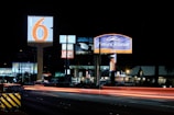 A nighttime scene featuring illuminated signs for various businesses including Motel 6 and Howard Johnson Inn, with light trails from passing vehicles on the road. Several other business signs are visible in the background.
