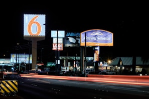 A nighttime scene featuring illuminated signs for various businesses including Motel 6 and Howard Johnson Inn, with light trails from passing vehicles on the road. Several other business signs are visible in the background.
