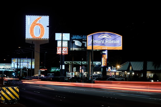 A nighttime scene featuring illuminated signs for various businesses including Motel 6 and Howard Johnson Inn, with light trails from passing vehicles on the road. Several other business signs are visible in the background.