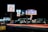 A nighttime scene featuring illuminated signs for various businesses including Motel 6 and Howard Johnson Inn, with light trails from passing vehicles on the road. Several other business signs are visible in the background.