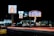 A nighttime scene featuring illuminated signs for various businesses including Motel 6 and Howard Johnson Inn, with light trails from passing vehicles on the road. Several other business signs are visible in the background.