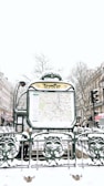 A snowy urban scene featuring a Paris Métro entrance named Temple. The entrance displays a map and is surrounded by ornate green metalwork with snow covering various surfaces. In the background, there are buildings lined with traditional Parisian architecture, bare winter trees, and multiple people walking by, dressed in winter clothing.