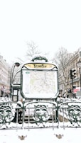 A snowy urban scene featuring a Paris Métro entrance named Temple. The entrance displays a map and is surrounded by ornate green metalwork with snow covering various surfaces. In the background, there are buildings lined with traditional Parisian architecture, bare winter trees, and multiple people walking by, dressed in winter clothing.