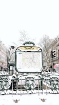 A snowy urban scene featuring a Paris Métro entrance named Temple. The entrance displays a map and is surrounded by ornate green metalwork with snow covering various surfaces. In the background, there are buildings lined with traditional Parisian architecture, bare winter trees, and multiple people walking by, dressed in winter clothing.