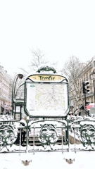 A snowy urban scene featuring a Paris Métro entrance named Temple. The entrance displays a map and is surrounded by ornate green metalwork with snow covering various surfaces. In the background, there are buildings lined with traditional Parisian architecture, bare winter trees, and multiple people walking by, dressed in winter clothing.