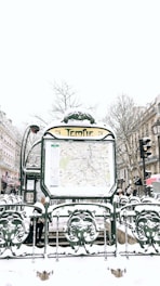 A snowy urban scene featuring a Paris Métro entrance named Temple. The entrance displays a map and is surrounded by ornate green metalwork with snow covering various surfaces. In the background, there are buildings lined with traditional Parisian architecture, bare winter trees, and multiple people walking by, dressed in winter clothing.