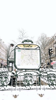 A snowy urban scene featuring a Paris Métro entrance named Temple. The entrance displays a map and is surrounded by ornate green metalwork with snow covering various surfaces. In the background, there are buildings lined with traditional Parisian architecture, bare winter trees, and multiple people walking by, dressed in winter clothing.