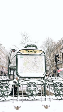 A snowy urban scene featuring a Paris Métro entrance named Temple. The entrance displays a map and is surrounded by ornate green metalwork with snow covering various surfaces. In the background, there are buildings lined with traditional Parisian architecture, bare winter trees, and multiple people walking by, dressed in winter clothing.
