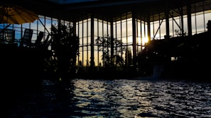 Indoor poolside view with silhouetted architecture and foliage as sunlight filters through large glass windows. Reflections ripple on the water surface, with chairs and an umbrella faintly visible to the left.