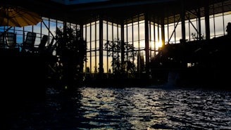 Indoor poolside view with silhouetted architecture and foliage as sunlight filters through large glass windows. Reflections ripple on the water surface, with chairs and an umbrella faintly visible to the left.