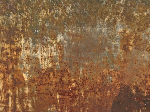 The image displays a heavily rusted surface with a mix of brown, orange, and yellowish tones. The texture is rough and uneven, indicating corrosion over time, with patches of peeling and worn-out metal.