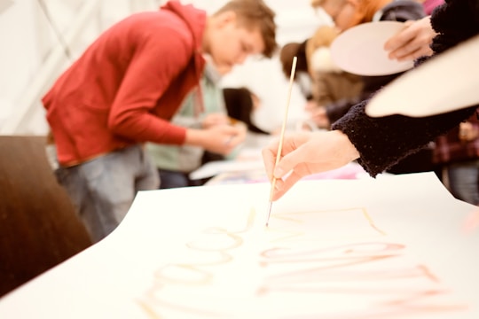 People are engaged in a creative activity, focusing on painting or drawing. One person is holding a paintbrush to a piece of paper. The background is blurred, suggesting a busy environment with multiple individuals participating.