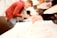 People are engaged in a creative activity, focusing on painting or drawing. One person is holding a paintbrush to a piece of paper. The background is blurred, suggesting a busy environment with multiple individuals participating.