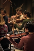 Musicians are intensely focused while playing a variety of string instruments, creating a harmonious atmosphere. The musician in the foreground is wearing headphones and holding a violin, contributing to a group rehearsal or performance setting.