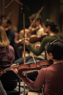 Musicians are intensely focused while playing a variety of string instruments, creating a harmonious atmosphere. The musician in the foreground is wearing headphones and holding a violin, contributing to a group rehearsal or performance setting.