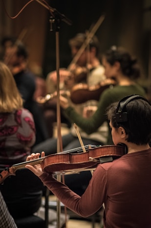 Musicians are intensely focused while playing a variety of string instruments, creating a harmonious atmosphere. The musician in the foreground is wearing headphones and holding a violin, contributing to a group rehearsal or performance setting.