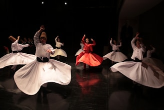 A group of people is performing a traditional dance with spinning motions. They are wearing flowing garments, primarily white, and one dancer in the center is dressed in red. They appear to be performing on a stage with dramatic lighting, creating a sense of motion and rhythm.
