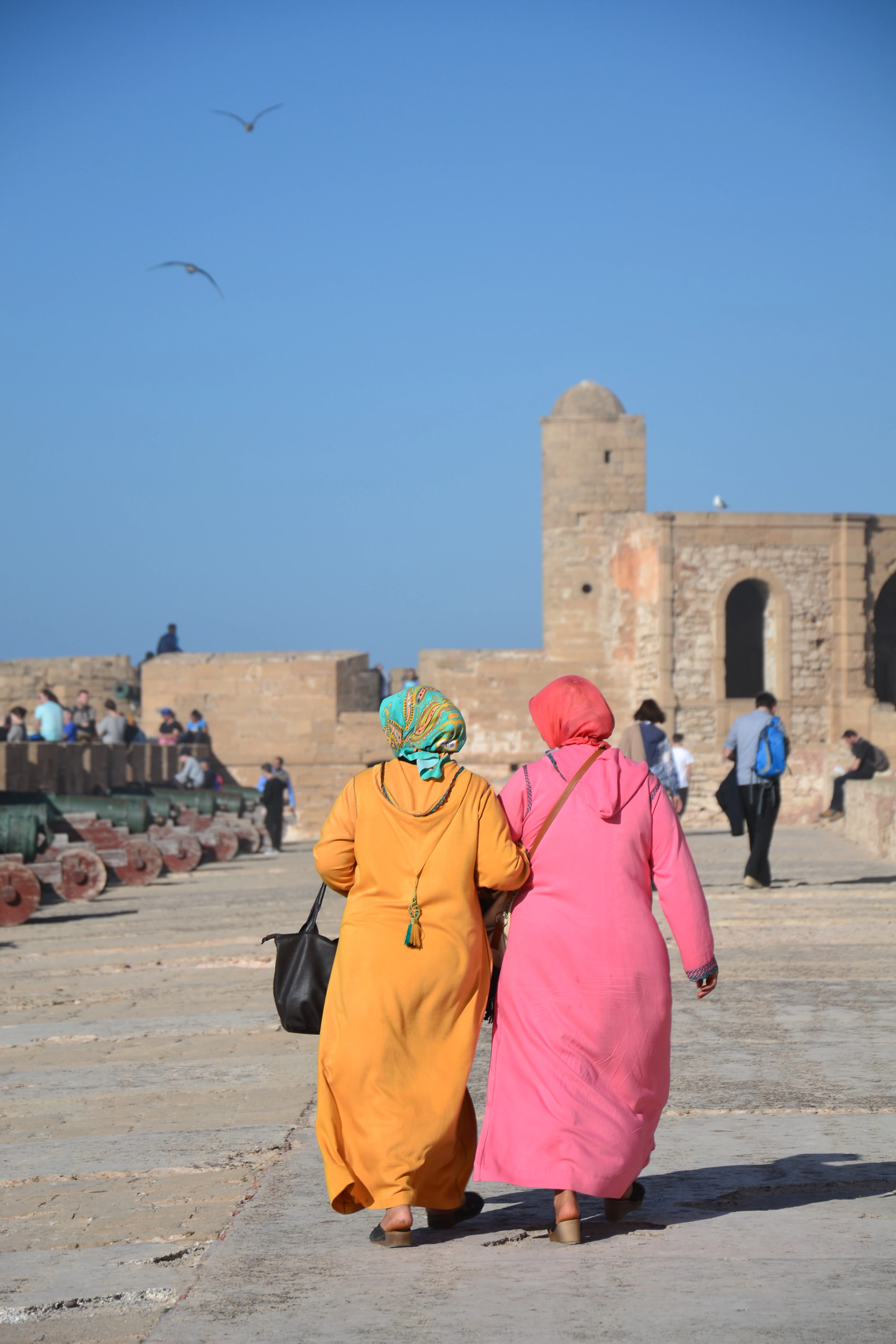 Women walking along a busy old-town street lined with shops and stone walls in Essaouira