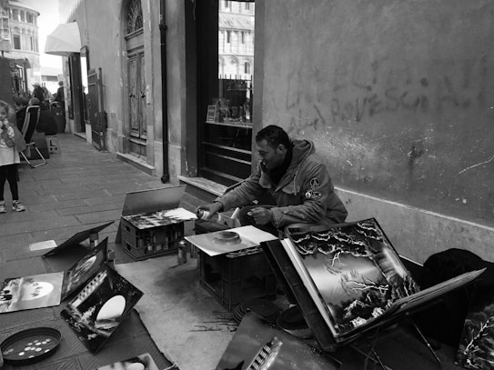 A street artist is seated on the pavement, surrounded by his paintings. He appears to be working on a new piece. The scene is monochromatic, with a few pedestrians and a child visible in the background, walking through an alleyway lined with old buildings. The artist has arranged his artworks and some spray paint cans around him.