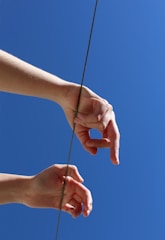 Two hands are positioned in mid-air against a clear blue sky, with a thin rope or wire running between them vertically.