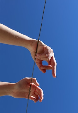 Two hands are positioned in mid-air against a clear blue sky, with a thin rope or wire running between them vertically.