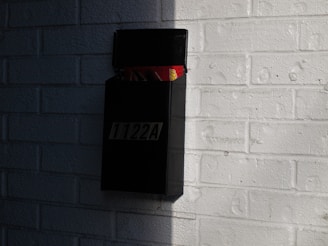 A black wall-mounted mailbox is attached to a white brick wall. The mailbox is open and contains envelopes and newspapers. Sunlight casts shadows on the wall, creating a contrast between light and dark areas.