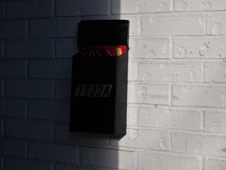 A black wall-mounted mailbox is attached to a white brick wall. The mailbox is open and contains envelopes and newspapers. Sunlight casts shadows on the wall, creating a contrast between light and dark areas.