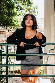 A person with short dark hair and bangs stands confidently behind a green metal barrier, wearing a black blazer over a black top and a light gray plaid skirt. Sunlight filters through the leaves of a tree in the background, casting dappled shadows on the ground. The scene is outdoors, set against a backdrop of greenery and an urban structure.