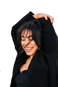 A woman with dark hair joyfully smiles while raising one arm above her head. She is wearing a black jacket and a black top, and stands against a plain white background.