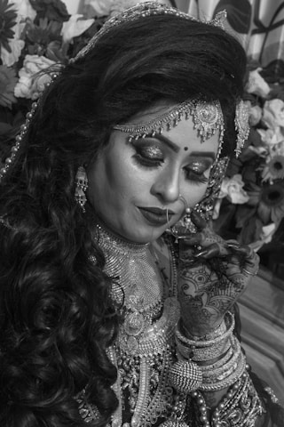 A woman wearing ornate traditional bridal attire and jewelry. Her eyes are closed, and her hands are adorned with intricate henna designs. She wears an elaborate headpiece and a nose ring. The background includes floral decorations.