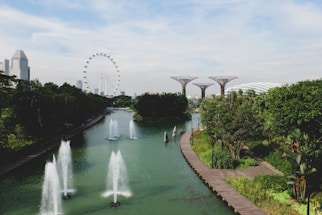 A scenic view of a lush garden with multiple tall, unique tree-like structures and a body of water with fountains in the foreground. In the background, an observation wheel and modern architecture blend with surrounding greenery, creating a balance between nature and urban design.
