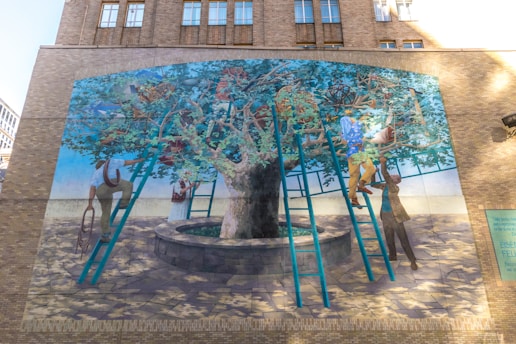 A mural on a brick wall depicts people interacting with a large tree, using ladders to reach its branches. The tree is situated in a circular stone planter. Various objects, including scientific instruments and books, are amidst the foliage. The scene conveys a sense of exploration and curiosity.