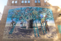 A mural on a brick wall depicts people interacting with a large tree, using ladders to reach its branches. The tree is situated in a circular stone planter. Various objects, including scientific instruments and books, are amidst the foliage. The scene conveys a sense of exploration and curiosity.