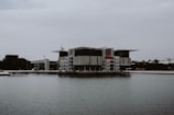 A large, modern building with industrial architecture sits by the waterfront. The structure features a rectangular design with prominent use of concrete and glass. Scaffolding and red accents are visible on the sides, and the building is surrounded by water, giving it an isolated appearance. Overcast skies create a subdued atmosphere.