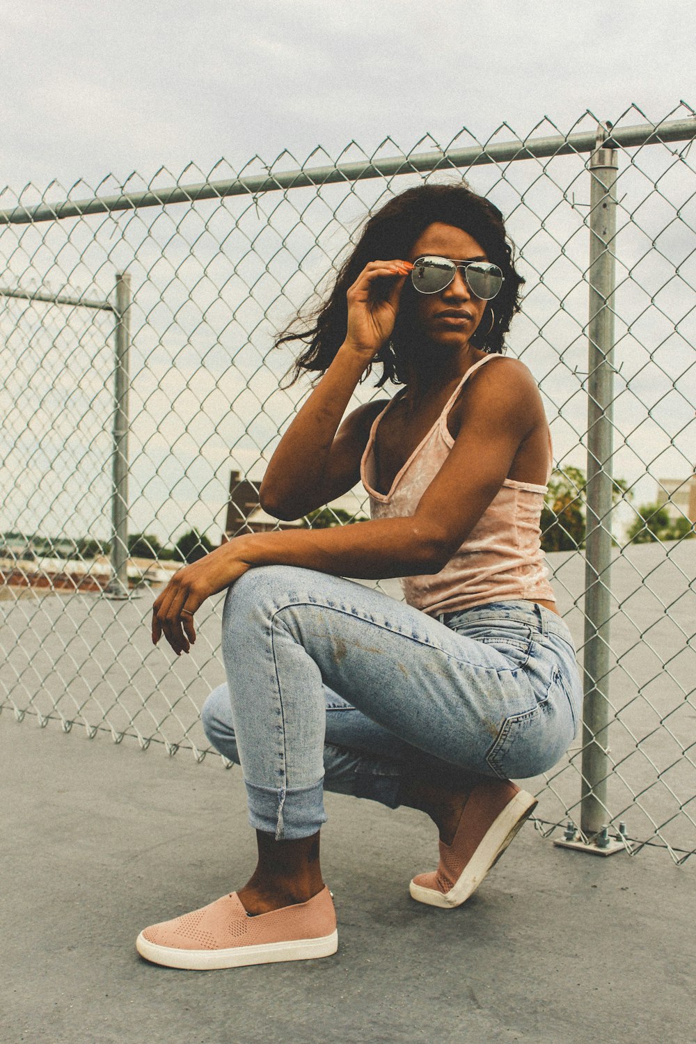 woman holding sunglasses while kneeling beside cyclone fence