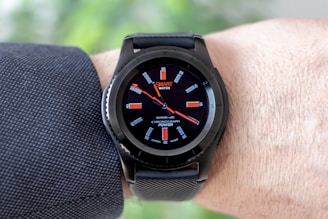 A black smartwatch with an analog-style display is worn on a person's wrist. It features a black strap and a round face with red and white detailing on the dial, showing both the brand and additional text. The wearer has a light skin tone and is dressed in a dark, textured fabric, possibly a suit or shirt.