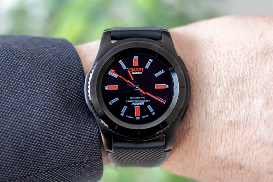 A black smartwatch with an analog-style display is worn on a person's wrist. It features a black strap and a round face with red and white detailing on the dial, showing both the brand and additional text. The wearer has a light skin tone and is dressed in a dark, textured fabric, possibly a suit or shirt.