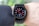 A black smartwatch with an analog-style display is worn on a person's wrist. It features a black strap and a round face with red and white detailing on the dial, showing both the brand and additional text. The wearer has a light skin tone and is dressed in a dark, textured fabric, possibly a suit or shirt.