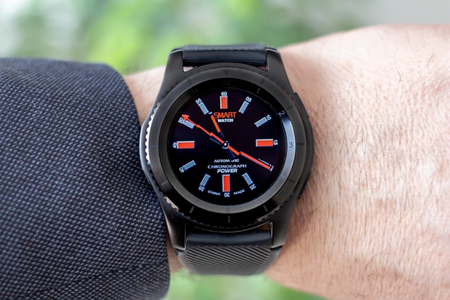 A black smartwatch with an analog-style display is worn on a person's wrist. It features a black strap and a round face with red and white detailing on the dial, showing both the brand and additional text. The wearer has a light skin tone and is dressed in a dark, textured fabric, possibly a suit or shirt.
