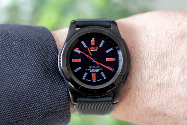 A black smartwatch with an analog-style display is worn on a person's wrist. It features a black strap and a round face with red and white detailing on the dial, showing both the brand and additional text. The wearer has a light skin tone and is dressed in a dark, textured fabric, possibly a suit or shirt.