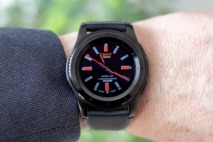 A black smartwatch with an analog-style display is worn on a person's wrist. It features a black strap and a round face with red and white detailing on the dial, showing both the brand and additional text. The wearer has a light skin tone and is dressed in a dark, textured fabric, possibly a suit or shirt.