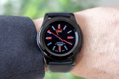 A black smartwatch with an analog-style display is worn on a person's wrist. It features a black strap and a round face with red and white detailing on the dial, showing both the brand and additional text. The wearer has a light skin tone and is dressed in a dark, textured fabric, possibly a suit or shirt.
